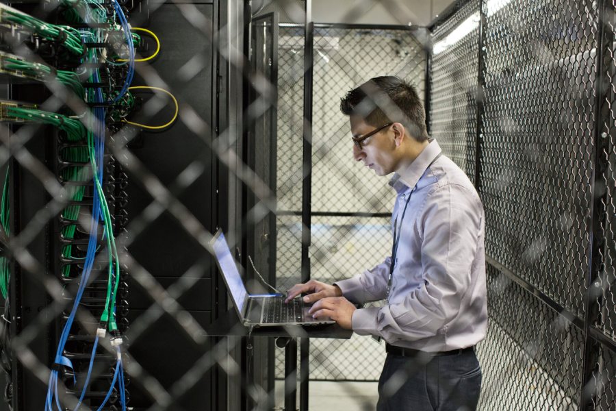Data center technician working on a laptop in a server room with network equipment and cables.