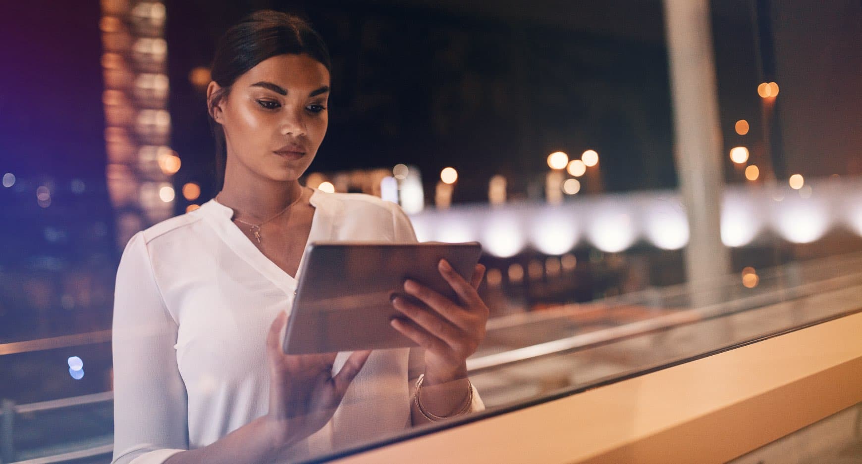 A young woman in white blouse using a tablet outdoors during nighttime with city lights in the background.