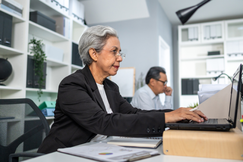 An elderly woman working on her laptop in a modern office setting.