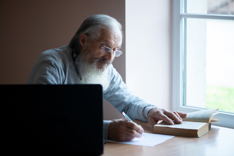 Elderly man reading and writing at home with a laptop and book, engaged in academic research and scholarly work.