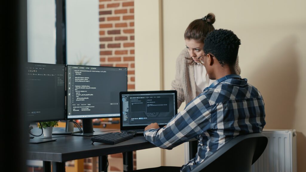 A woman and a man collaborate on coding projects using dual monitors and a laptop in a modern office setting.