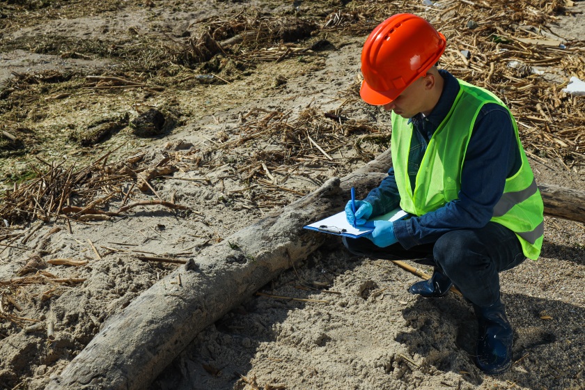 Alt: Investigator in safety gear examines beach debris after environmental disaster.