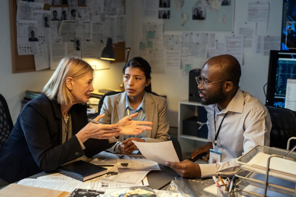 Photo of diverse colleagues engaged in a team meeting discussing research and academic papers in a busy office environment.