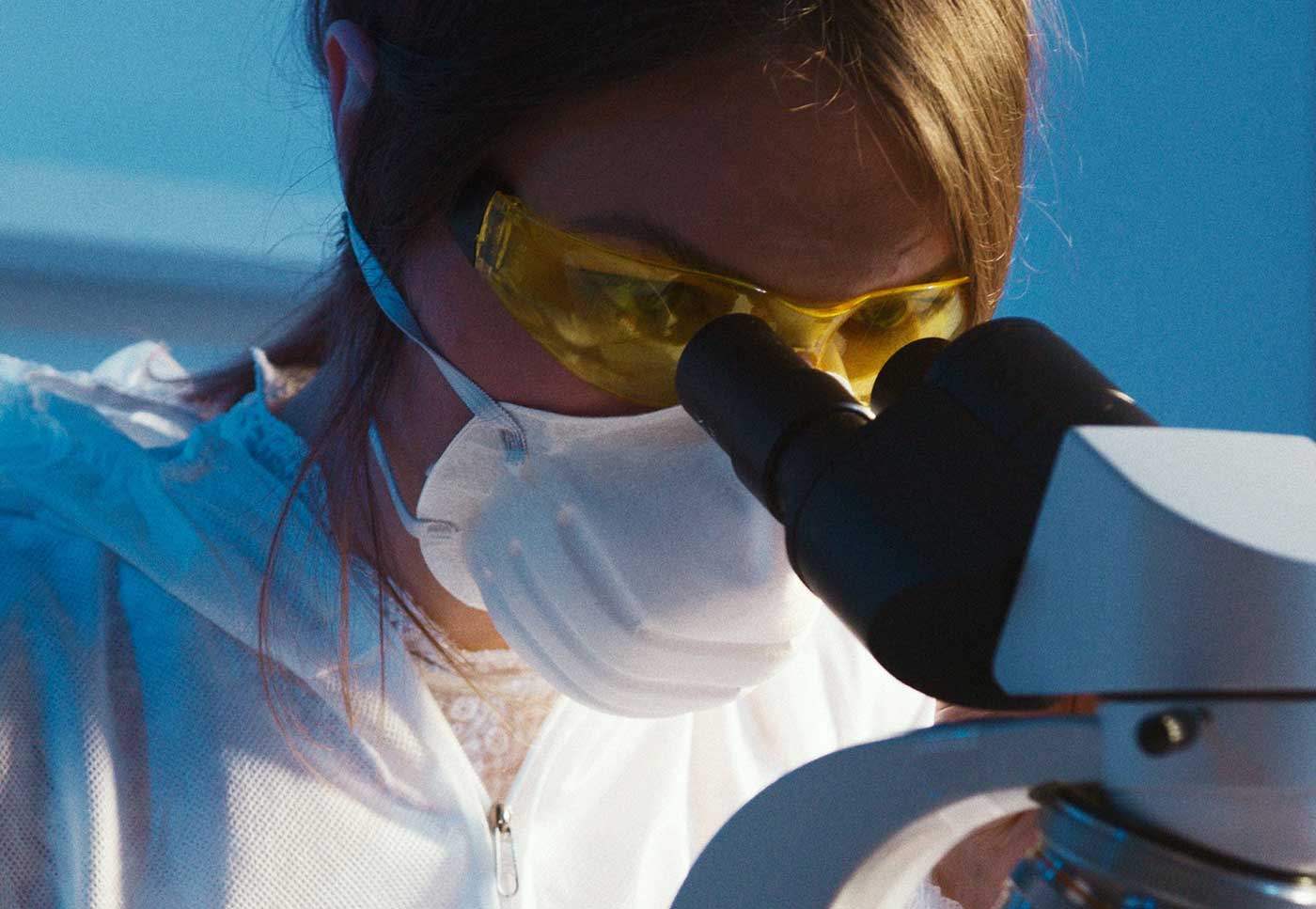 Close-up of a scientist using a microscope in a laboratory setting, wearing protective gear and safety glasses.