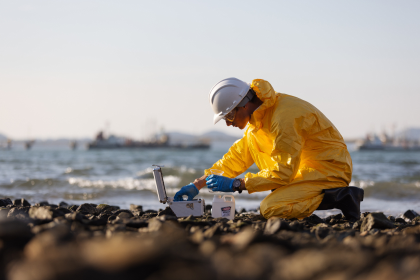Scientist in yellow protective suit collecting water samples at shoreline.