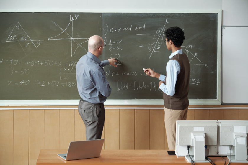 An instructor and student discussing complex mathematical equations on a classroom blackboard.