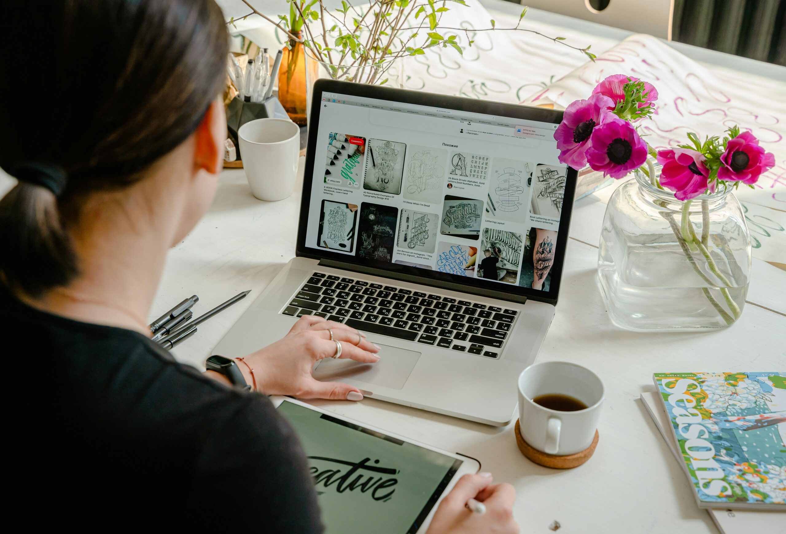An individual working on digital illustration and typography design on a laptop, surrounded by art supplies and a flower vase.