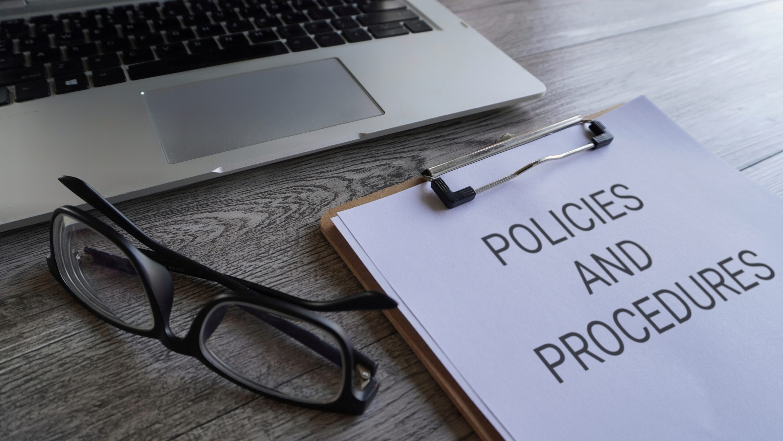 A policy document titled "POLICIES AND PROCEDURES" on a clipboard with glasses and a laptop.
