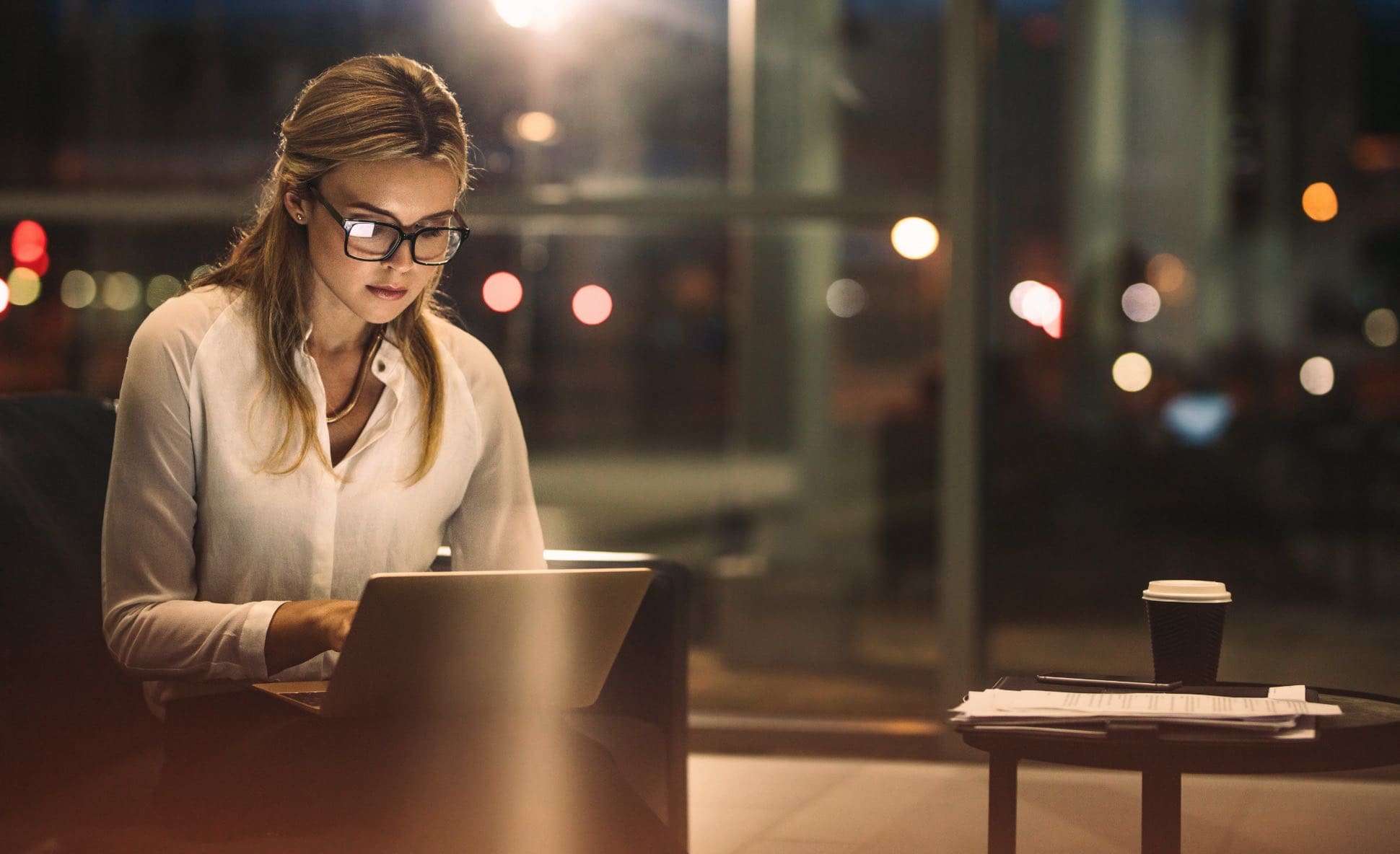 A professional woman working on a laptop in a modern, dimly lit setting with a coffee cup and documents nearby.