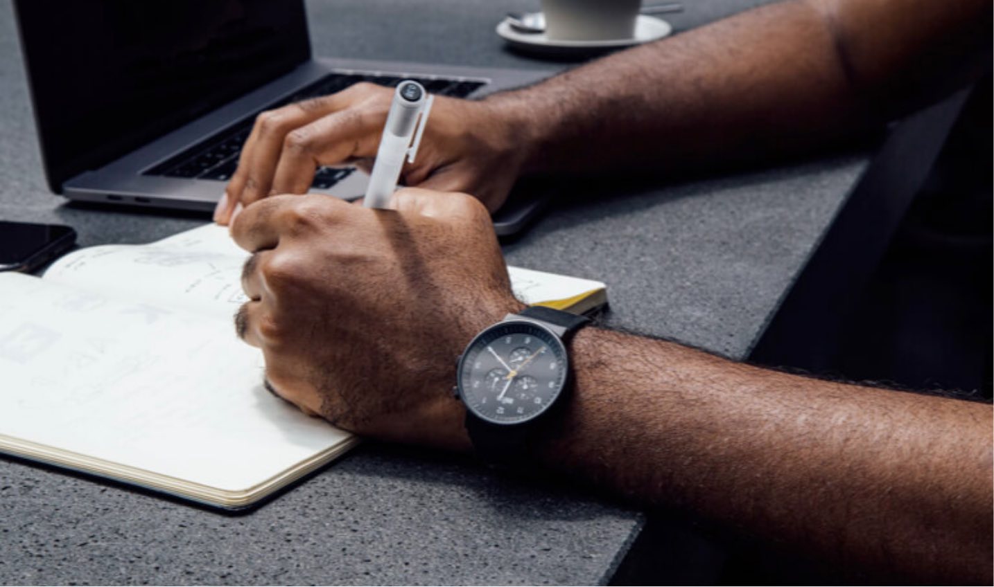 Modern workspace with a person writing in a notebook, using a pen, with a laptop nearby for academic research.