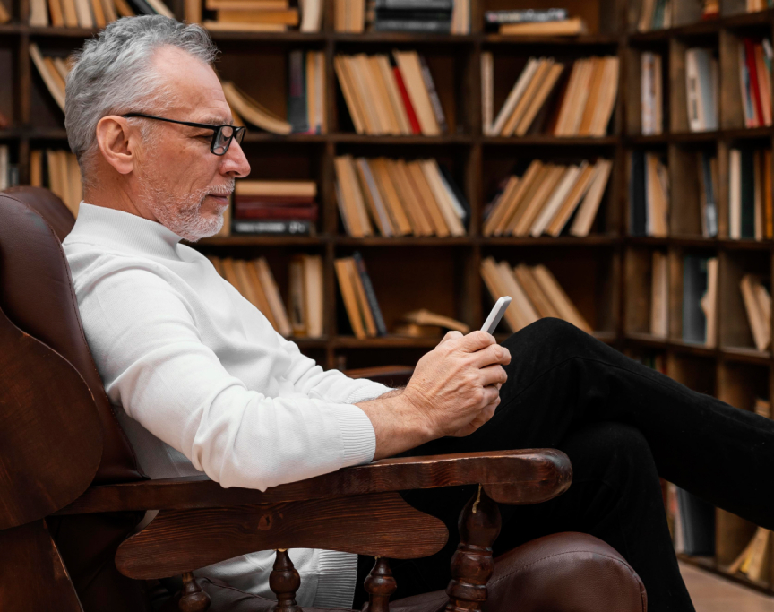 An academic man in a library using a smartphone, surrounded by shelves of books.