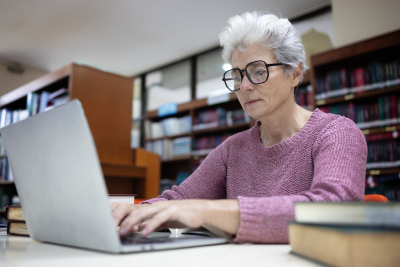 Older woman using laptop in a library setting for academic research on global journals.