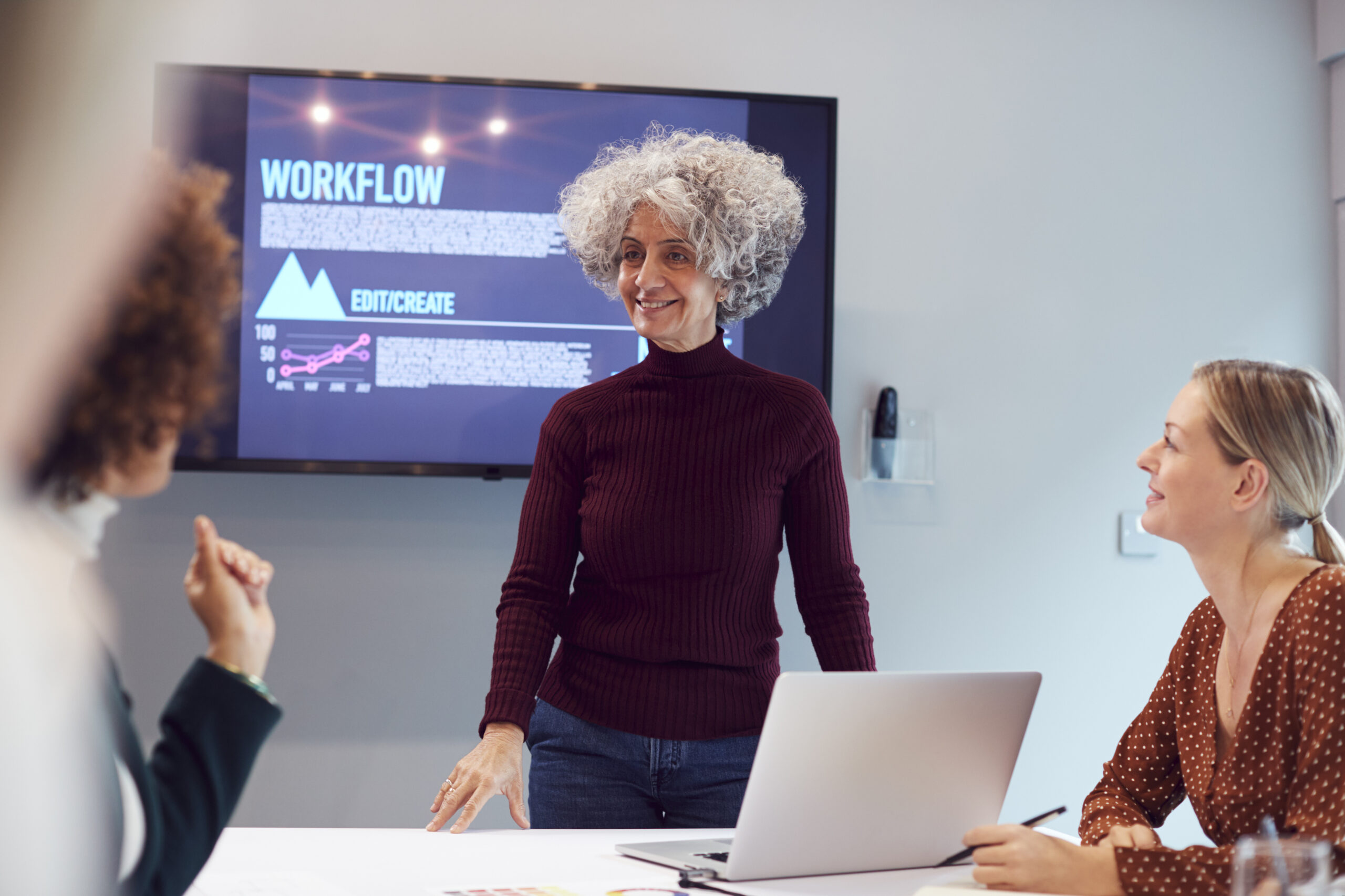 Diverse group of women collaborating in an academic research meeting discussing workflow processes with presentation screen in the background.